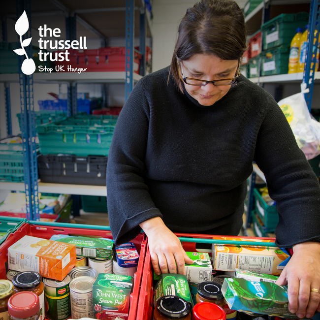 The logo of Trussell Trust is in the top left corner. A woman is helping assemble plastic trays filled with tins and food.